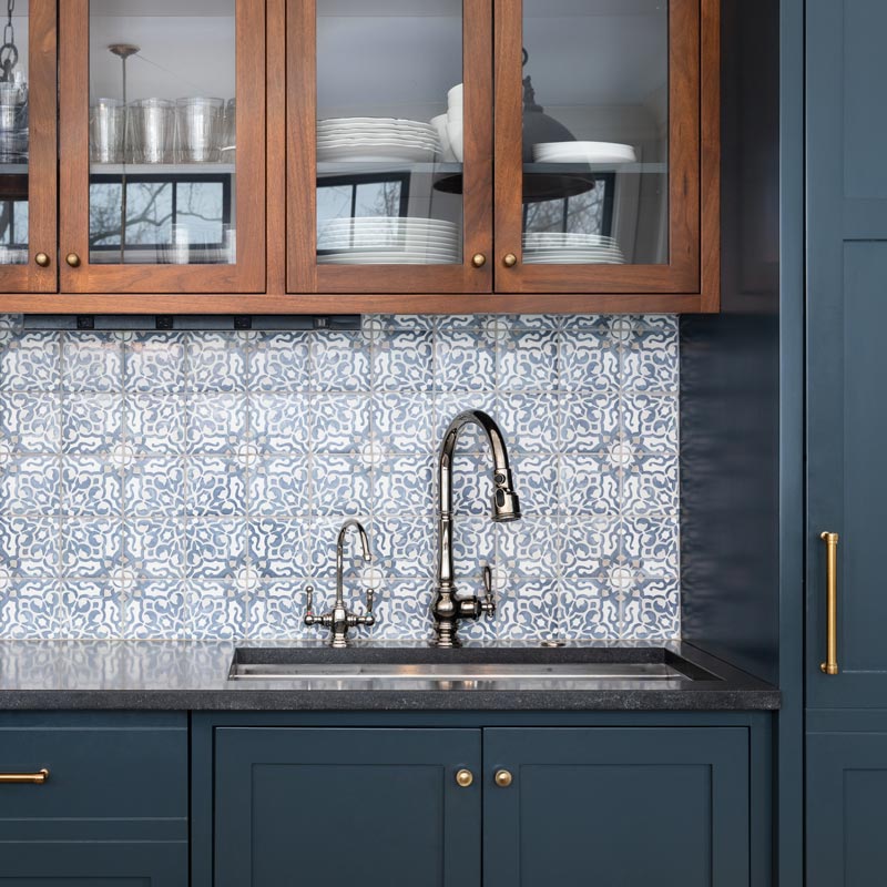 A kitchen sink with a beautiful pattern tiled backsplash with a chrome faucet, black granite countertops, and surrounded by blue and wood cabinets.